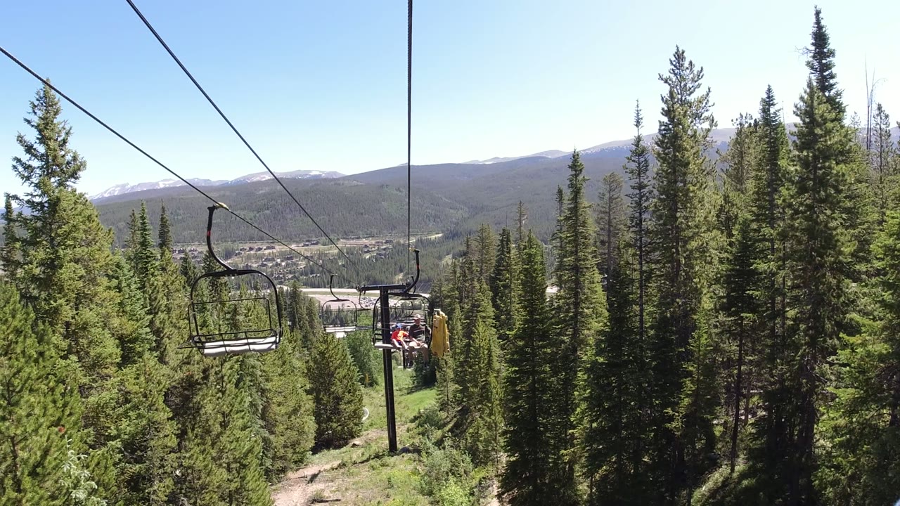 Winter Park, Colorado lift cables run diagonally across the sky, with a black chair ahead, tall evergreens filling the slope, distant hills, and no visible people.