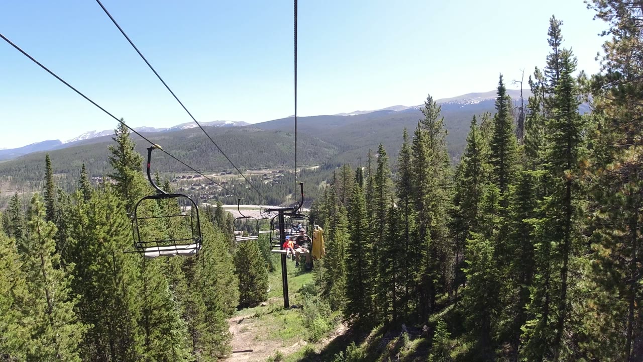 Winter Park, Colorado lift chairs hang along diagonal cables above dense pines, with seated riders visible in the center distance and hazy mountain slopes beyond.
