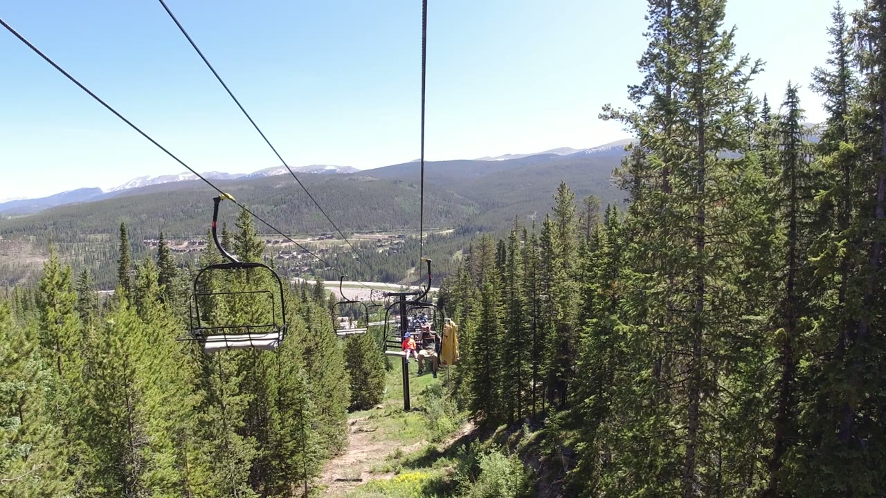 Winter Park, Colorado ski lift chairs hang over a narrow cleared path through dense evergreens, with mountain ridges in the background and no visible people.