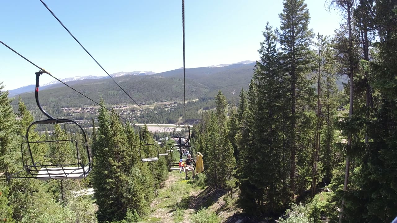 Opening frame from Winter Park, Colorado showing an empty chairlift seat on the left, diagonal lift cable, dense green trees, distant valley, and no visible people in this frame.