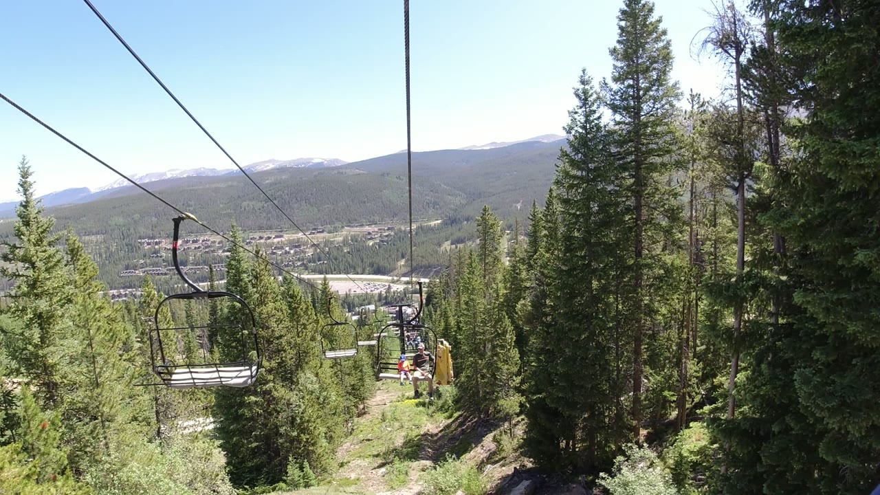 Winter Park, Colorado chairlift view with cables crossing the sky, an empty chair near center left, tall conifers framing the slope, and small riders visible farther down the line.