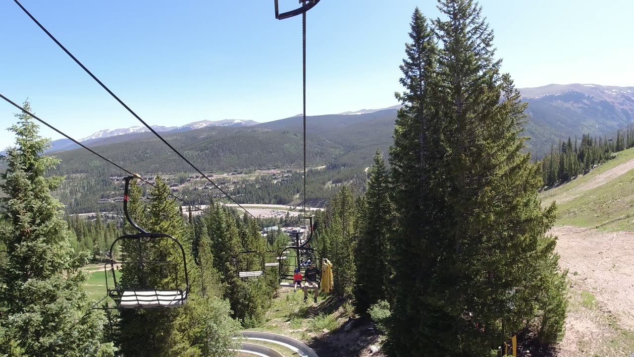 A chairlift cable runs diagonally past an empty chair near the center-left, with tall evergreens, a pale dirt run, distant ridges, and no visible people in Winter Park, Colorado.