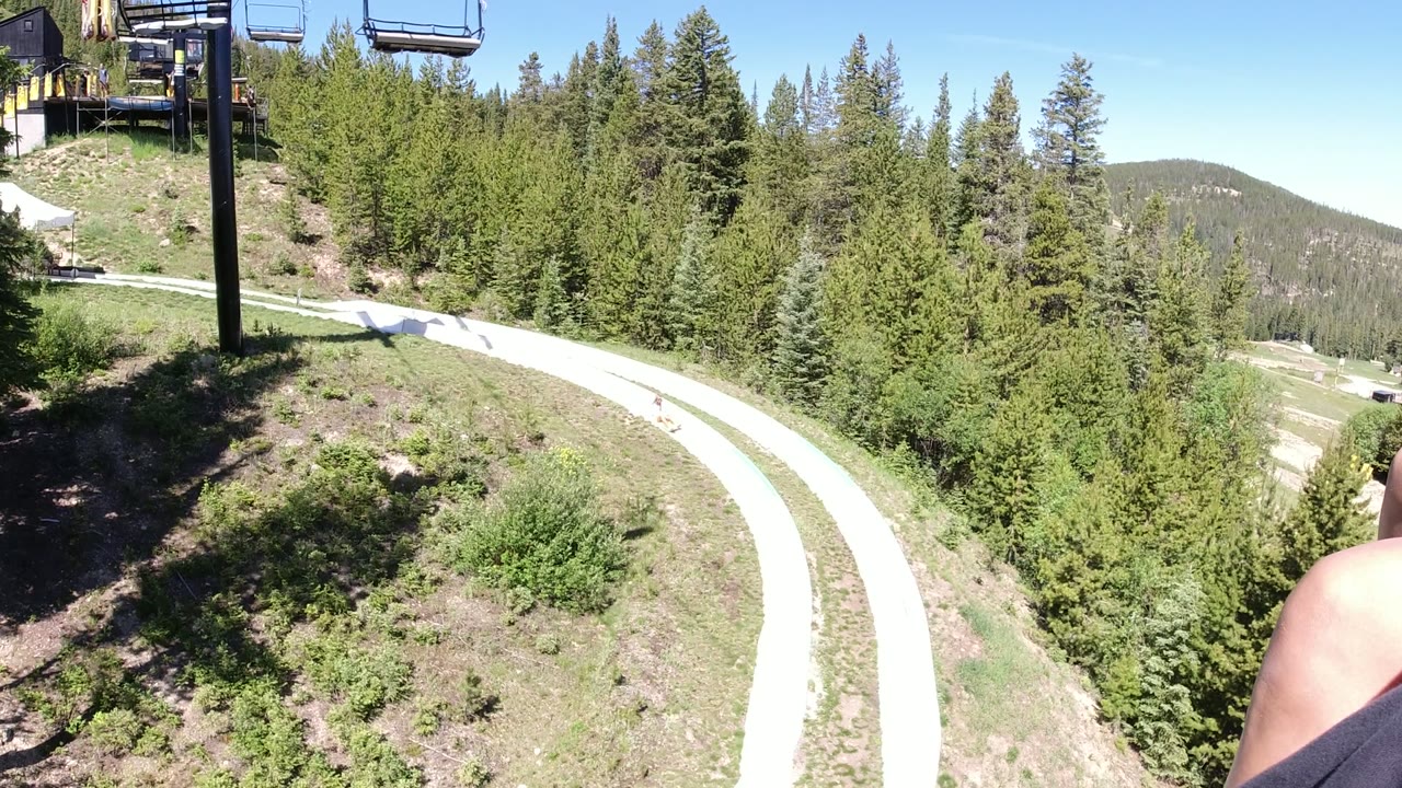 One second into the Winter Park chairlift ride, an empty lift chair hangs near the top of frame while the winding path and green trees stretch below.