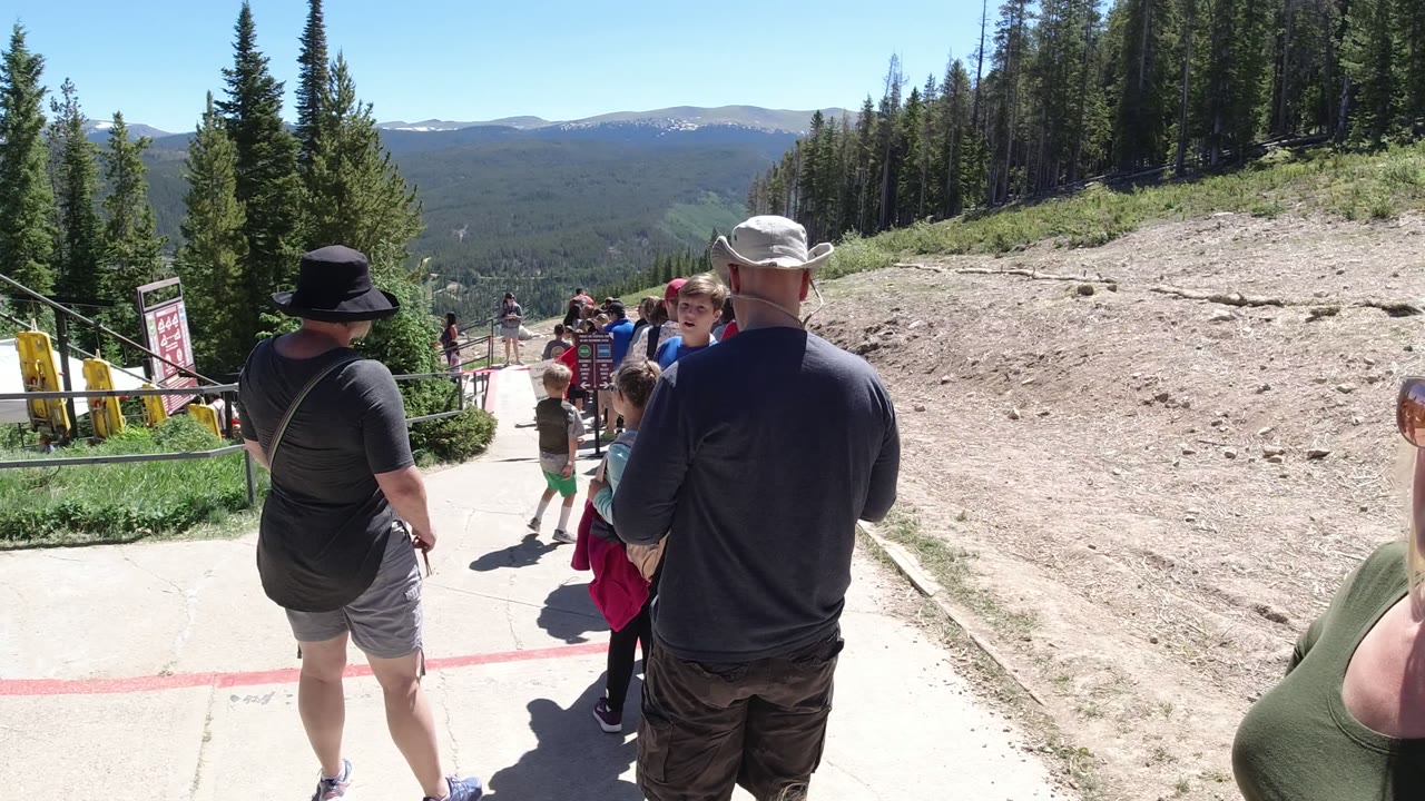 A group of visitors at Winter Park, Colorado stands in line near yellow railings and signs, with a child moving in the walkway and tree-covered ridges beyond.
