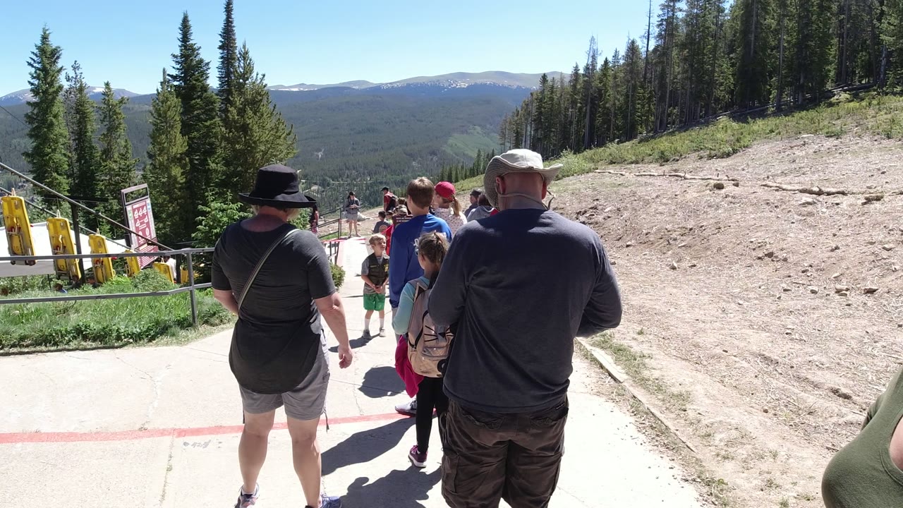 At 0:01 in Winter Park, Colorado, the line remains centered as a man in a dark hat, children, and other visitors face toward the rope area below the trees.