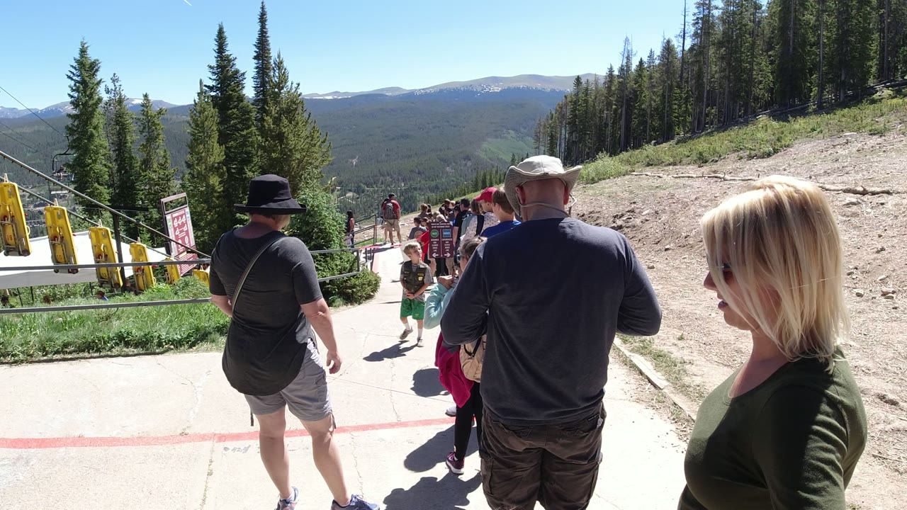 Tourists standing in line on a mountain walkway with pine trees and distant ridges.