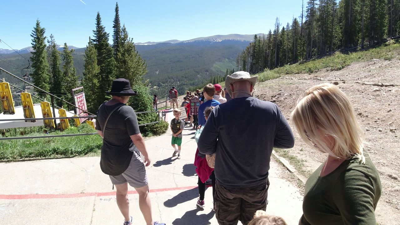 At 0:01 in Winter Park, Colorado, the crowd remains gathered along the walkway, with a blonde woman in the right foreground and a forested valley beyond the railings.