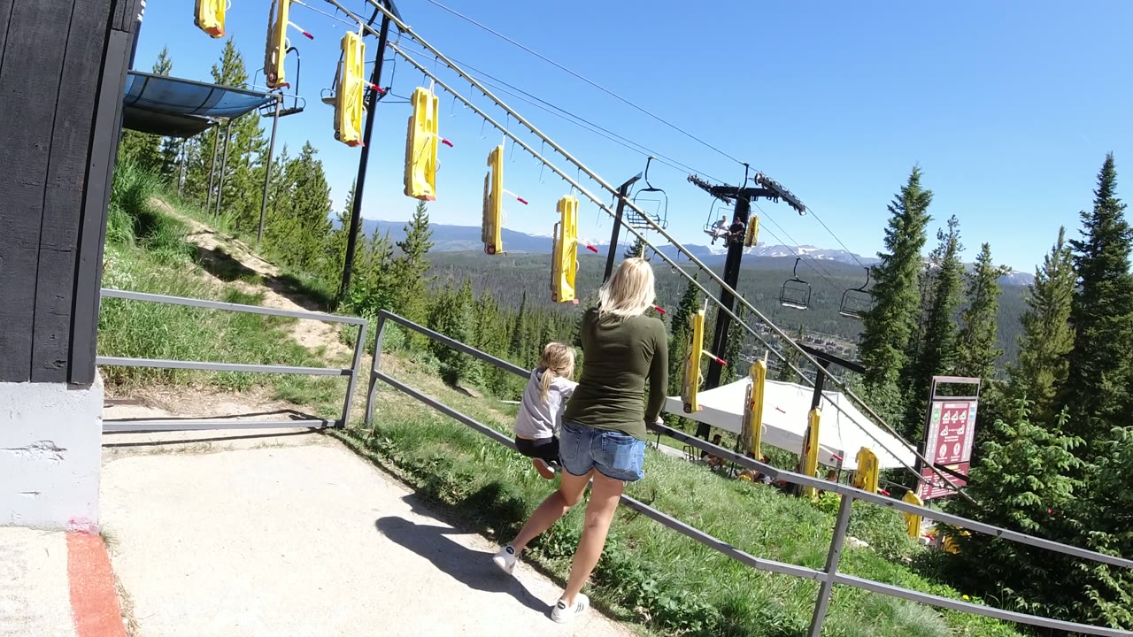 At 0:01 in Winter Park, Colorado, the woman and child shift slightly along the walkway, with yellow lift hangers overhead and a chairlift line descending behind them.