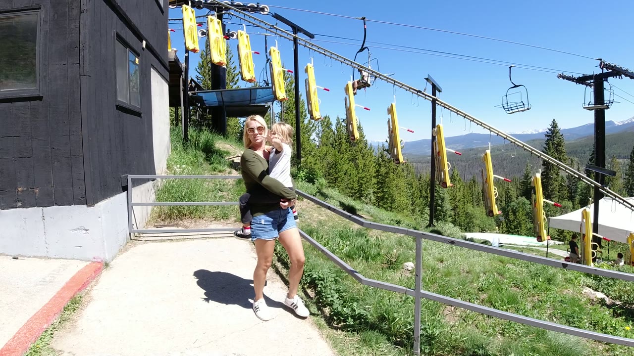 A woman holding a child faces the camera near a chairlift loading area in Winter Park, Colorado, with yellow hanging pads and forested slopes behind them.