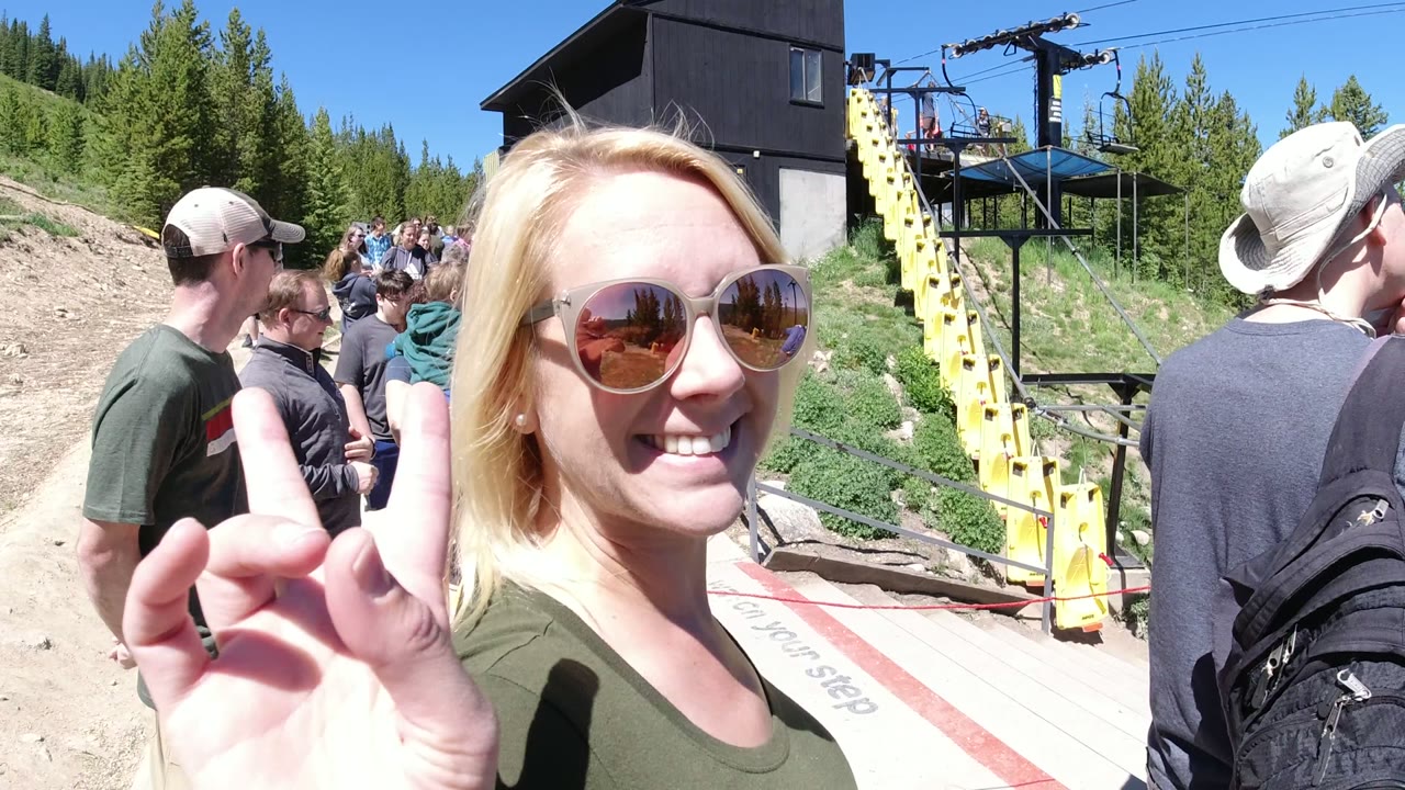 Blonde woman smiles at the camera and raises a peace sign in Winter Park, Colorado, while tourists wait near the yellow lift structure and dark building.