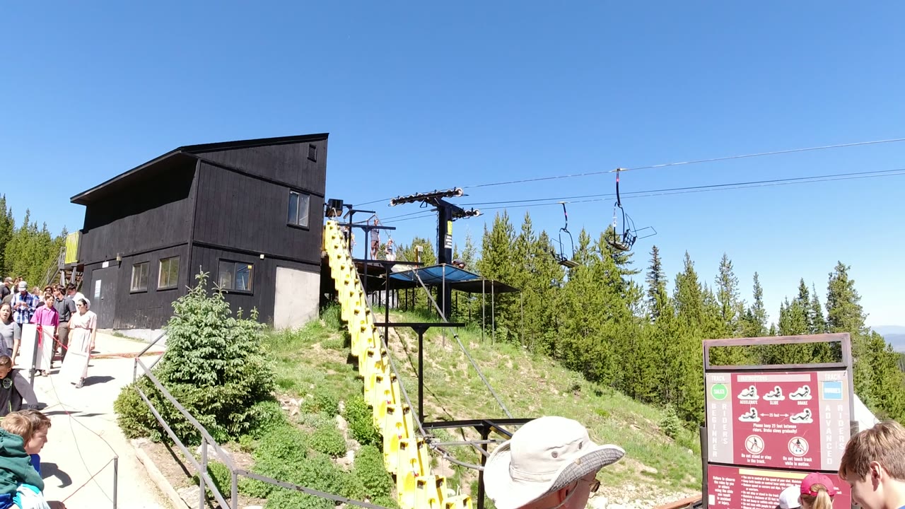 At Winter Park, Colorado, visitors stand and walk near a dark lift building, yellow chairlift support, red sign, evergreen trees, and clear blue sky.