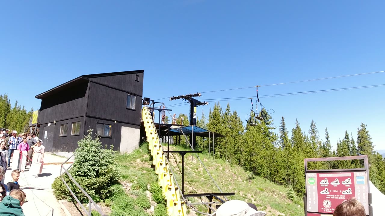 At Winter Park, Colorado, a group of visitors gathers along the walkway beside a dark lift building, visible lift cables, empty chairs, and green trees.