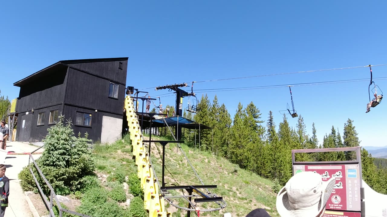 Wide frame of a Winter Park, Colorado lift attraction with a black building, yellow support tower, chairlift cables, pine trees, red signage, and visitors at the walkway edge.