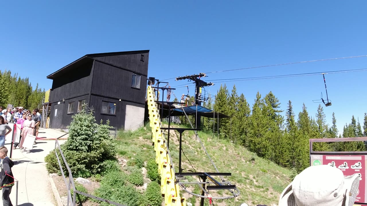 Winter Park, Colorado lift platform scene with a group of visitors on the left walkway, a black building above the slope, chairlift equipment, pine trees, and clear blue sky.