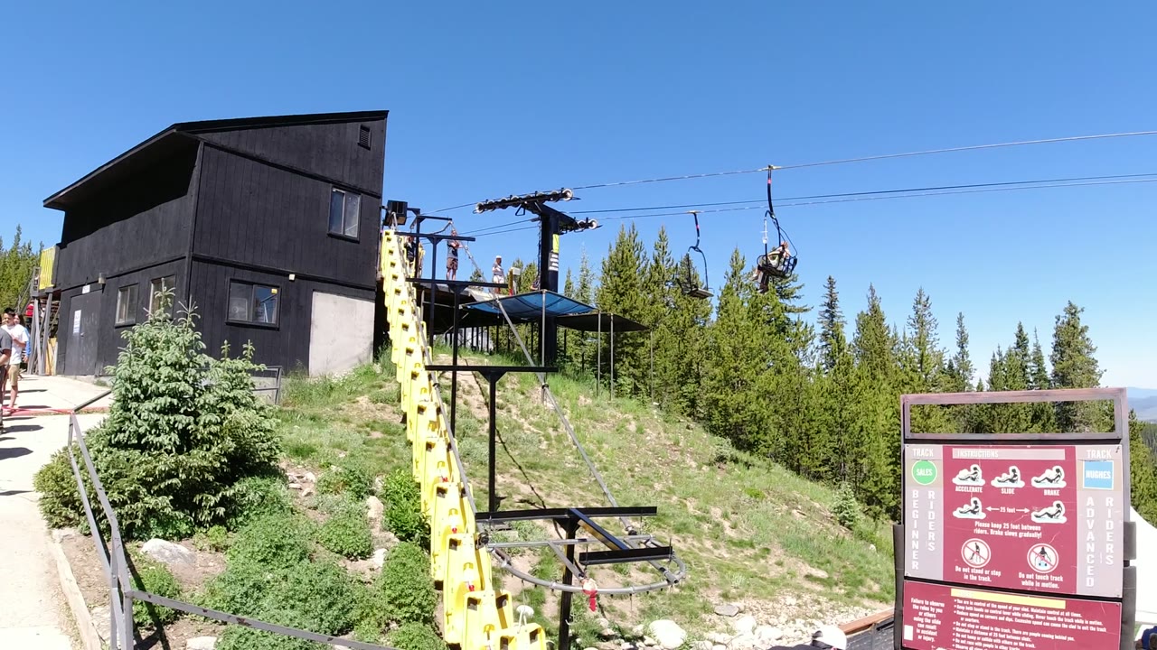 Winter Park, Colorado hilltop lift area with the black station building, yellow support tower, moving chairlift seats, conifer trees, red sign, and visitors partly visible near the walkway.