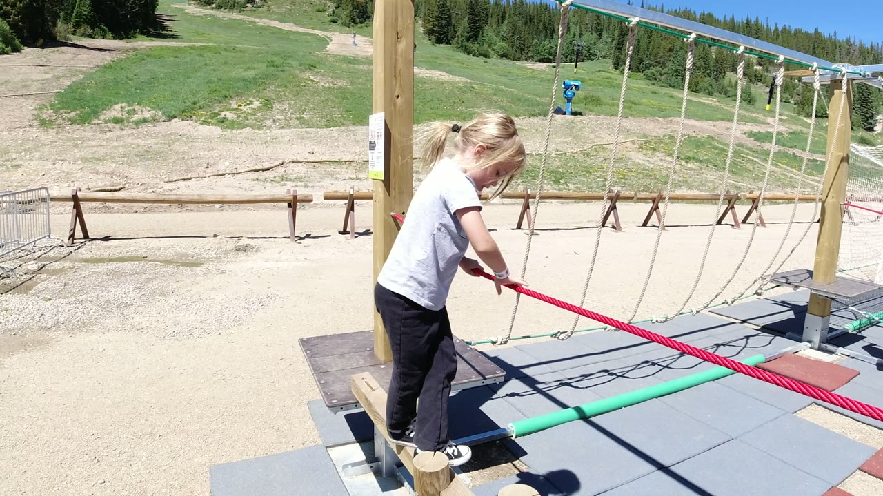 A blonde girl in Winter Park, Colorado holds a red rope while standing on a small wooden obstacle platform, with hanging ropes and grassy terrain behind her.
