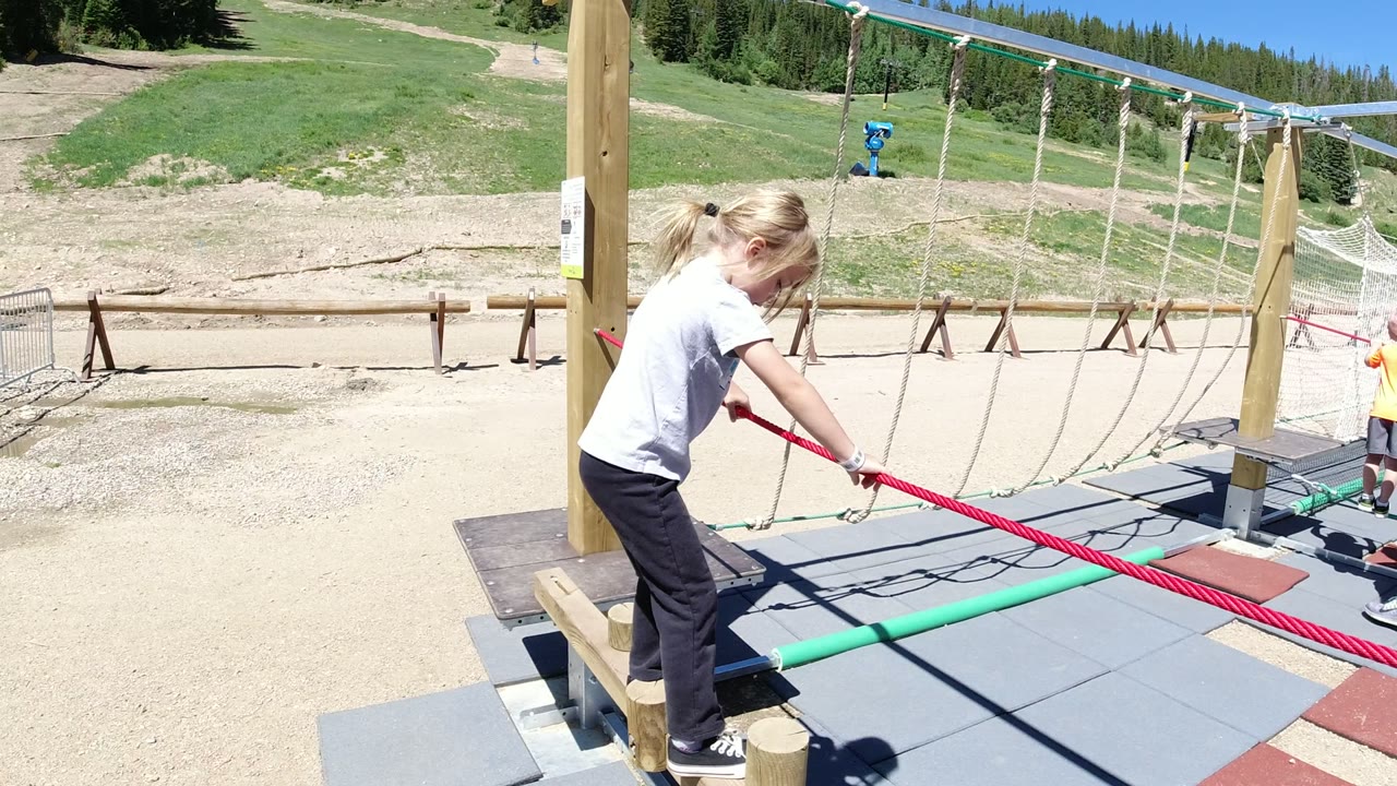 The girl leans forward on the Winter Park obstacle course, hands on the red rope, with a green balance pole crossing the padded platform below.