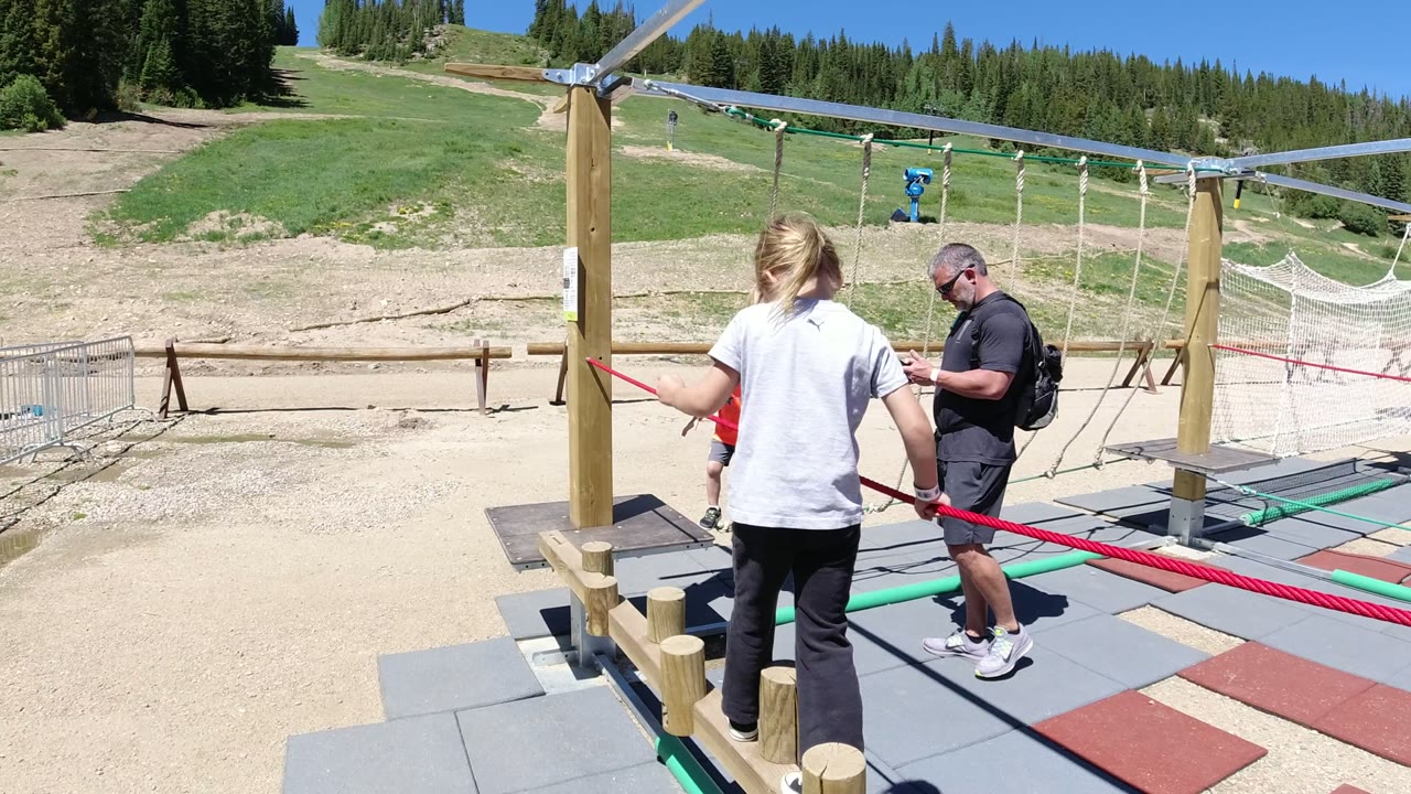 At 0:01 in Winter Park, Colorado, the girl stands closer to the red rope as an adult remains on the right side of the wood and rope course.