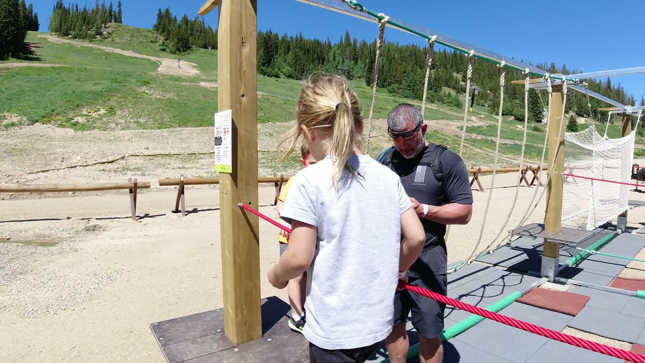 In Winter Park, Colorado, the girl stands with her back to camera on the rope-course platform while an adult man in dark clothing looks down at a phone.