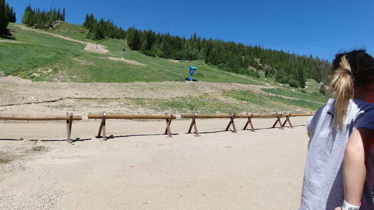 Wide ground-level view in Winter Park, Colorado showing sandy foreground, green mountain slopes, evergreen trees, blue sky, and a child partly visible at the right edge.
