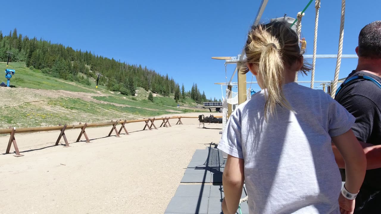 A young girl in a gray shirt walks toward a ropes course in Winter Park, Colorado, with sandy ground, wood fencing, green slopes, and evergreen trees behind her.