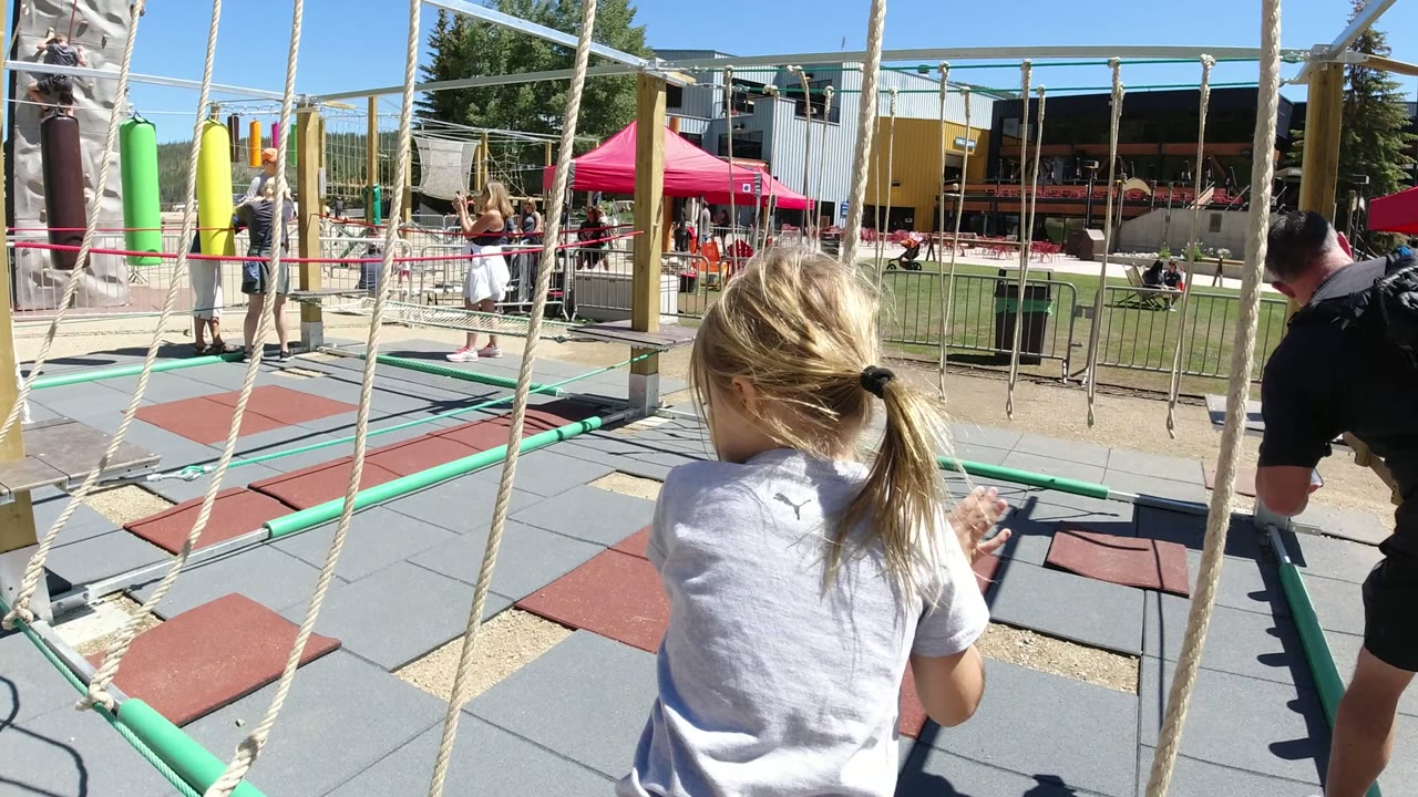 A young girl seen from behind approaches rope obstacles on an outdoor playground in Winter Park, Colorado, with adults and children beyond the railings.
