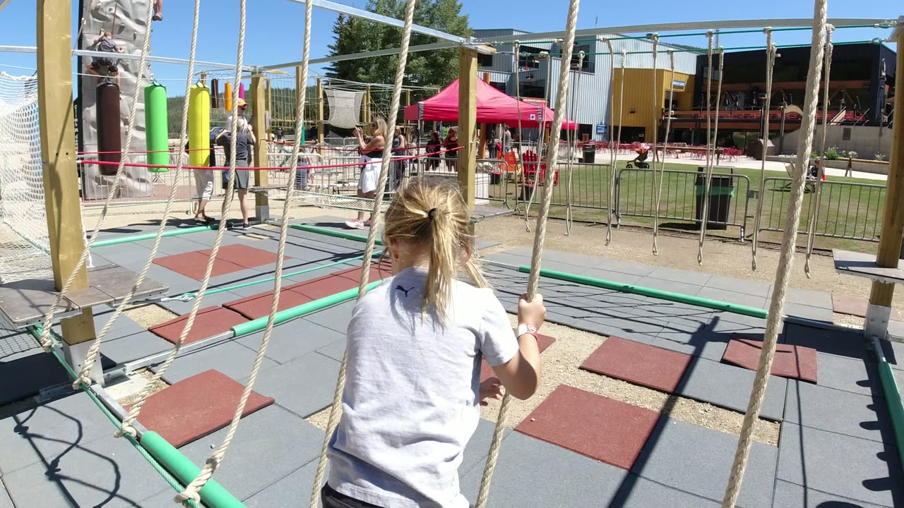 The girl grips a vertical rope on the playground course in Winter Park, Colorado, with red and gray safety tiles, tall posts, and people in the background.