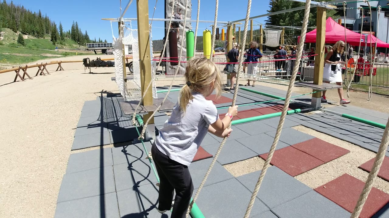 A young girl in Winter Park, Colorado holds two vertical ropes while stepping onto a low green rope over rubber safety tiles, with visitors and pine-covered slopes behind her.