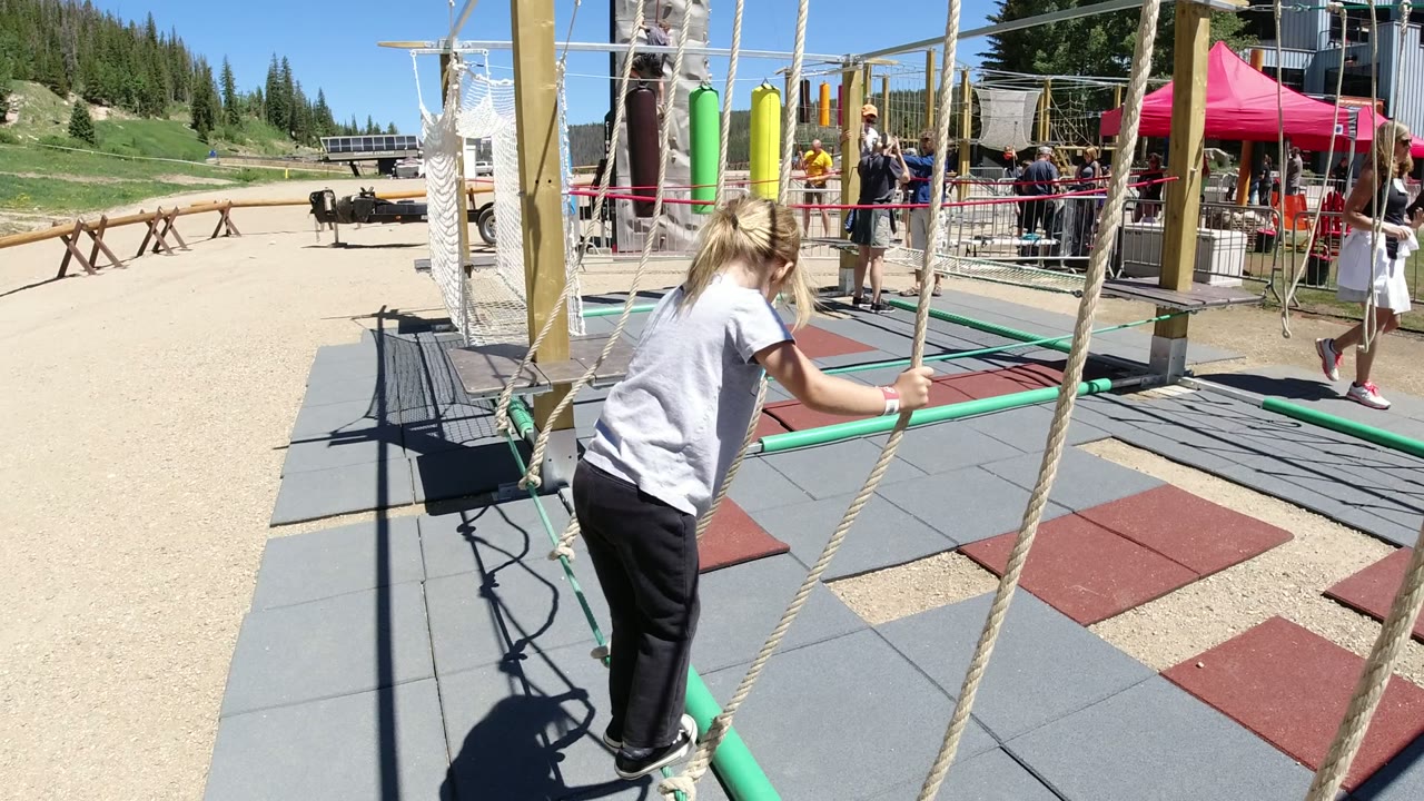 The girl leans forward on the green rope in Winter Park, Colorado, gripping the hanging ropes as rubber mats, posts, a red canopy, and other people fill the recreation area.