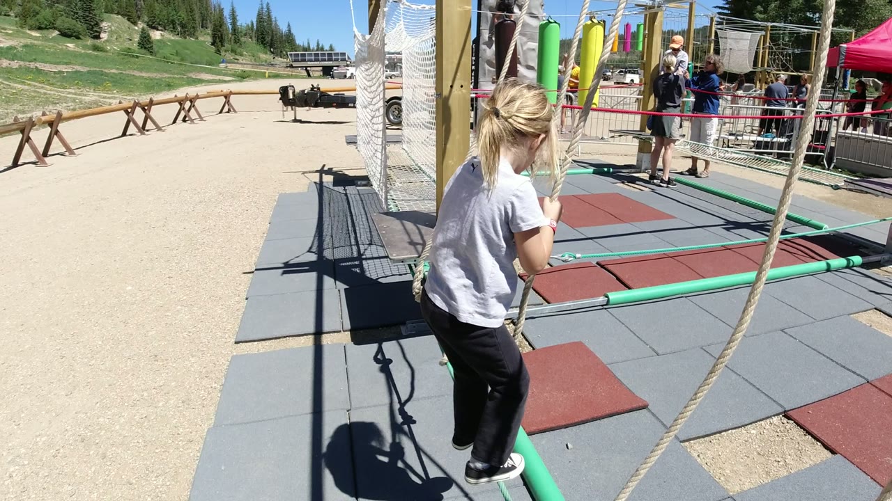The child stands on the green balance bar in Winter Park, Colorado while holding two ropes, with adults and a fenced activity area behind her.