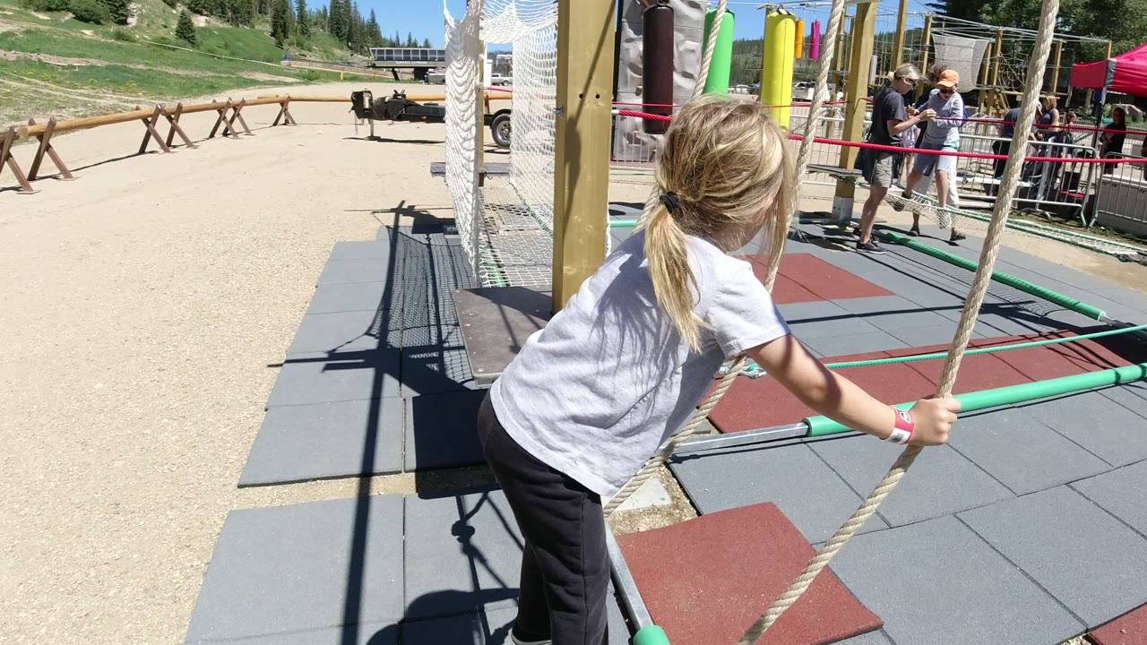 The girl leans farther forward on the rope swing lines in Winter Park, Colorado, with red and gray safety pads below and several adults gathered near the course railings.