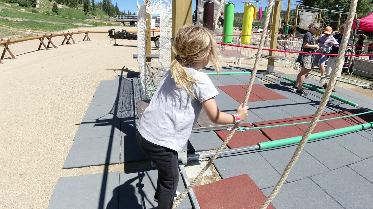 The girl leans forward on the Winter Park, Colorado rope course, gripping the hanging ropes as adults watch from behind the red barrier.