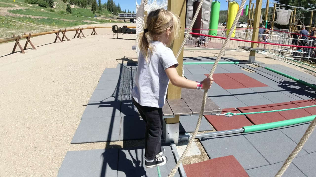 A young girl in Winter Park, Colorado stands near a thick rope and padded mats at the start of an outdoor ropes course, with fencing and mountain slope behind her.