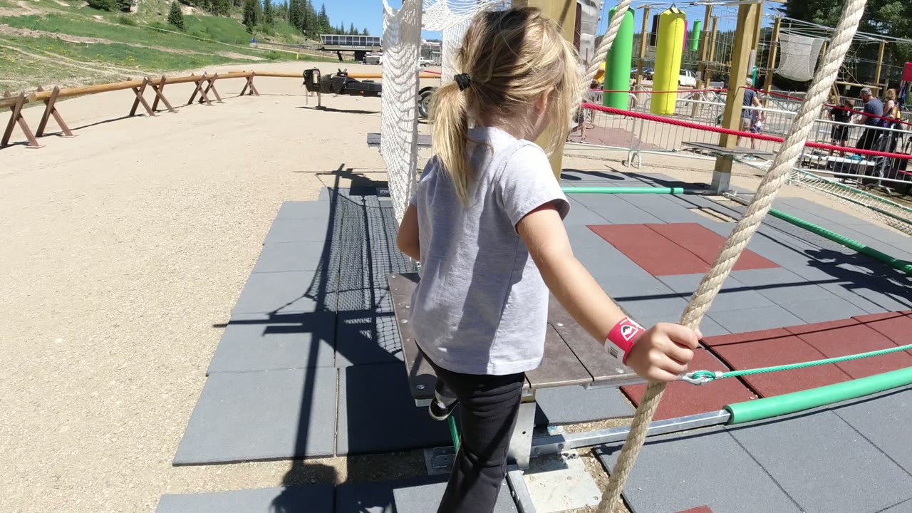 The child advances along the Winter Park, Colorado rope structure, holding the rope with one hand while padded gray and red mats fill the foreground.