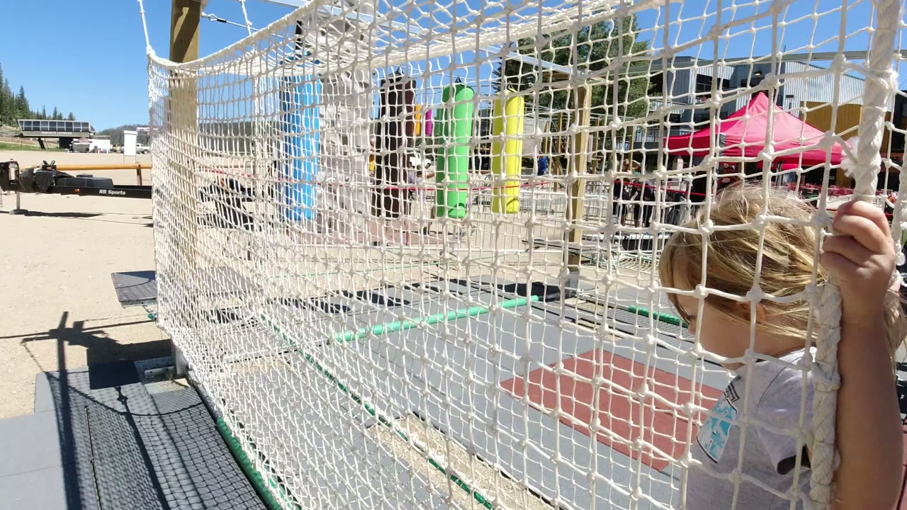 Young blonde girl partly hidden behind white rope netting at an outdoor play area.