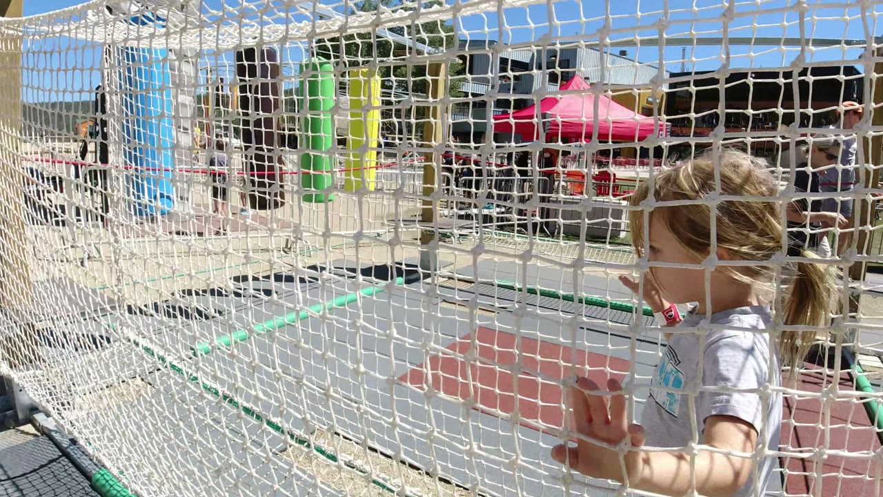 Blonde girl in a white shirt holds the rope net inside the Winter Park, Colorado play area, with green railings and colorful padding visible behind the mesh.