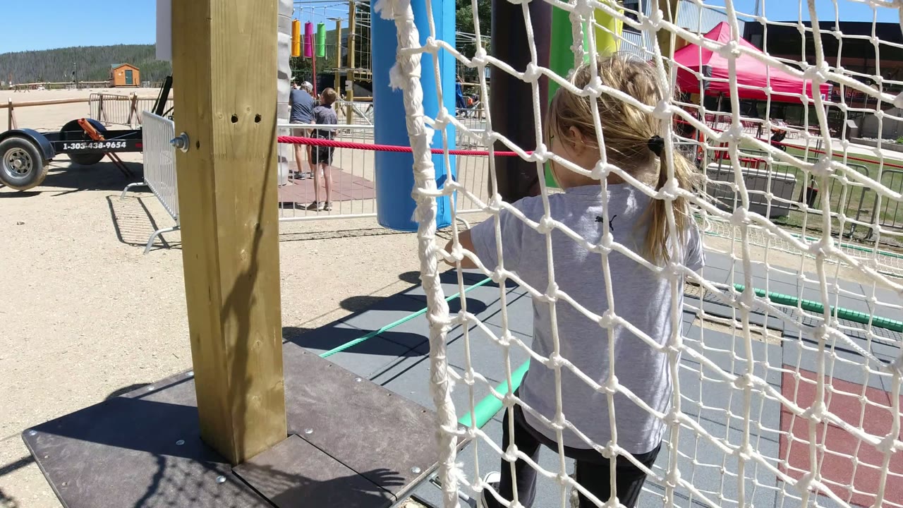 Child in Winter Park, Colorado stands close to the rope net, with blue and green padded posts, fencing, and a pink tent visible beyond the play area.