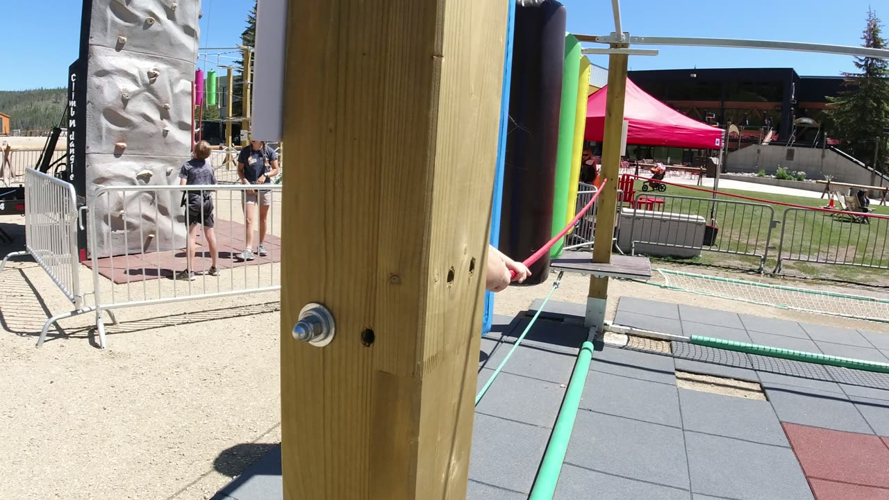 A Winter Park, Colorado playground rope course is seen close to a wooden support post, with a child's arm on a red rope and other people near a climbing wall in the background.