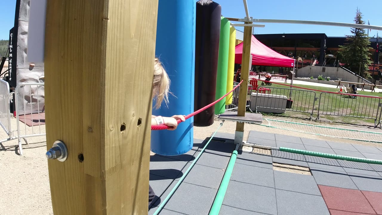 A blonde child leans into a red rope beside blue and black playground posts in Winter Park, Colorado, with railings, tents, grass, and distant people behind the play area.