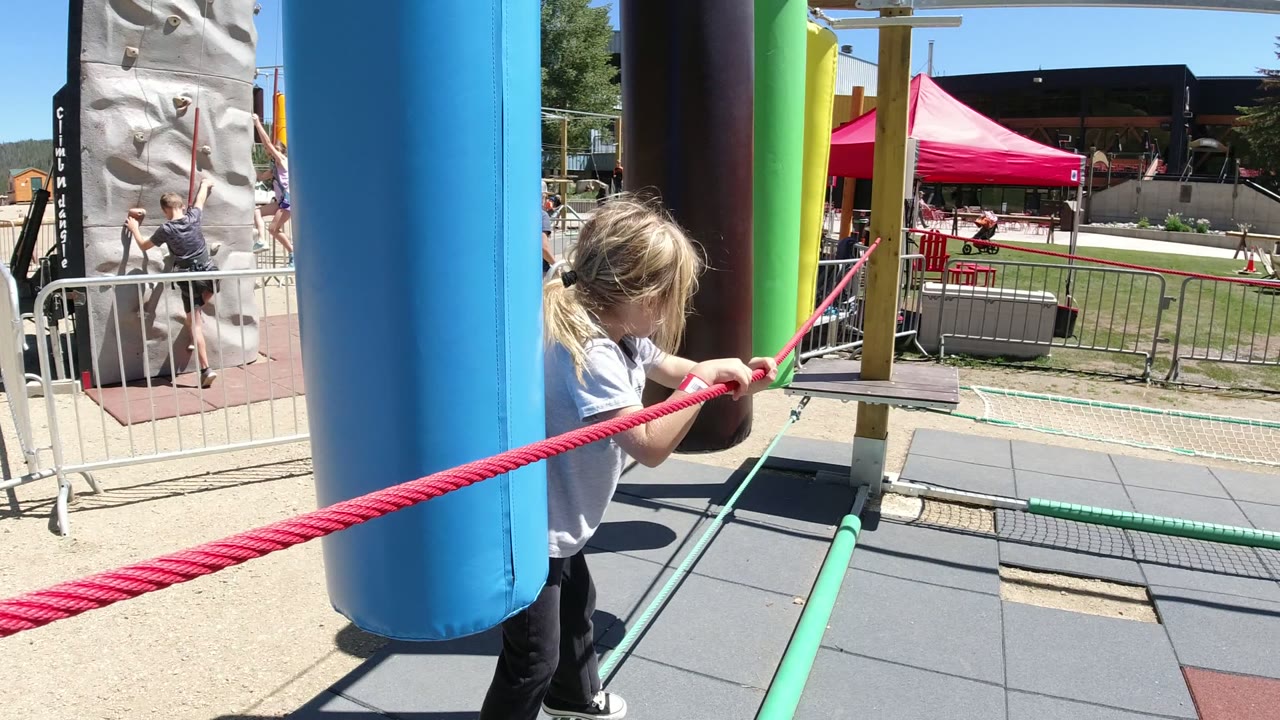A child in Winter Park, Colorado holds a red rope beside a blue padded obstacle post, with a climbing wall and metal barriers visible behind them.
