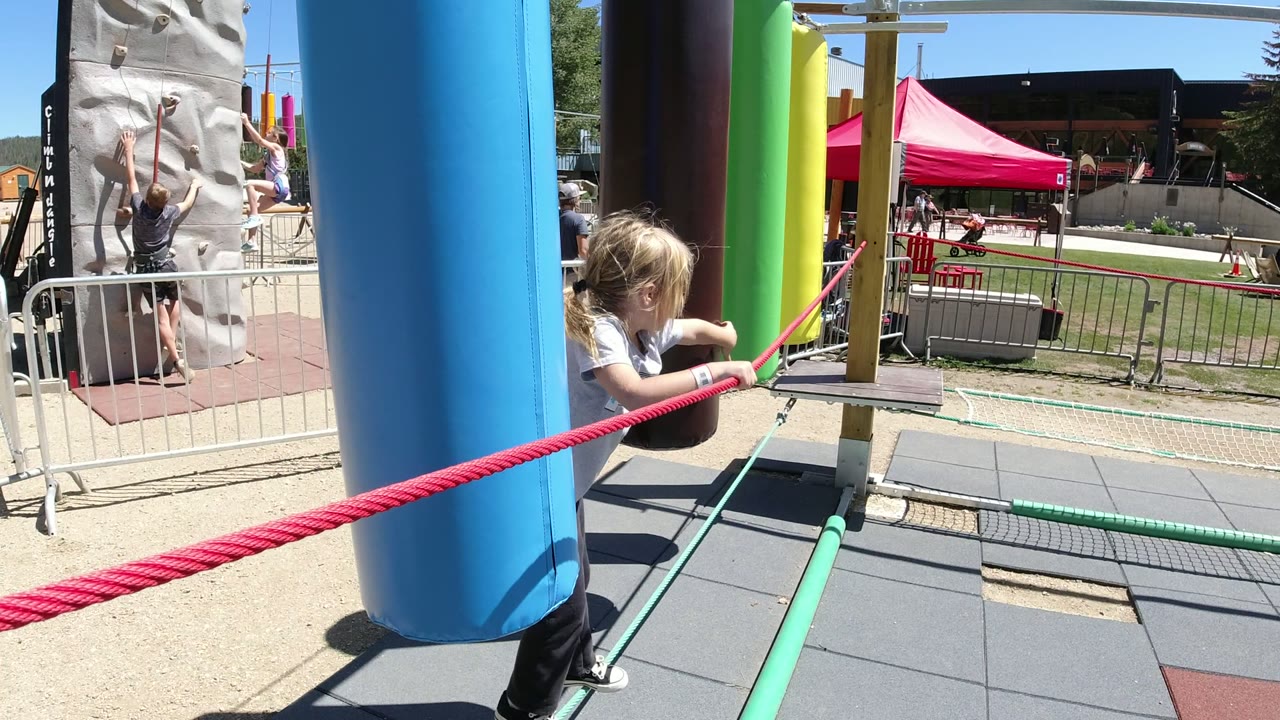 The child continues along the Winter Park, Colorado obstacle course, gripping the red rope while standing close to a large blue padded post.