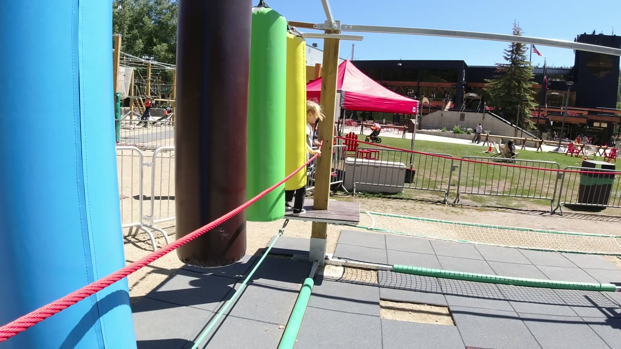 One second in, the young girl appears closer to the green and yellow padded posts on the Winter Park playground structure, with red and green ropes crossing the foreground and people seated in the background.