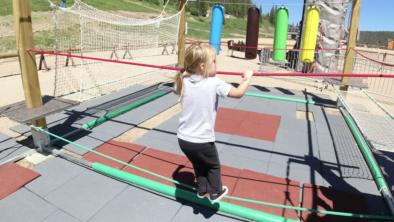 The girl continues across the rope course in Winter Park, Colorado, with her feet on a green rope and both hands near the red support rope.