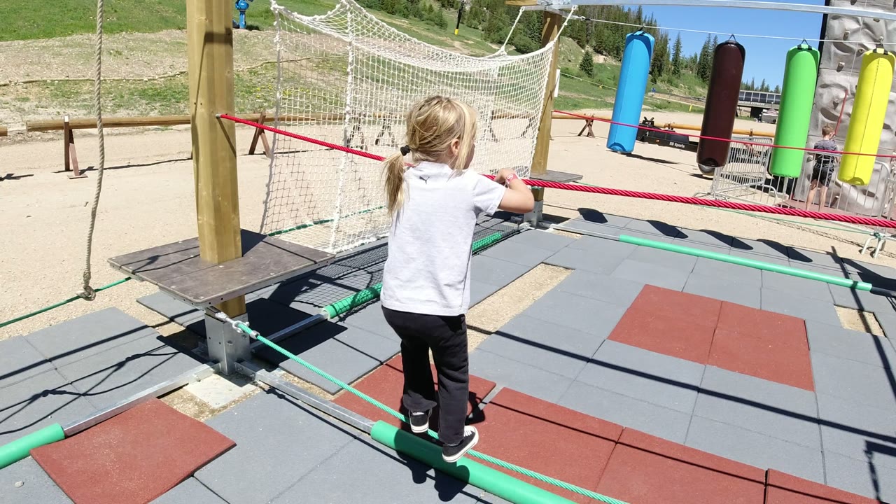 The girl shifts farther along the green rope path in Winter Park, Colorado, facing the netted play area while colorful hanging cylinders sit in the background.