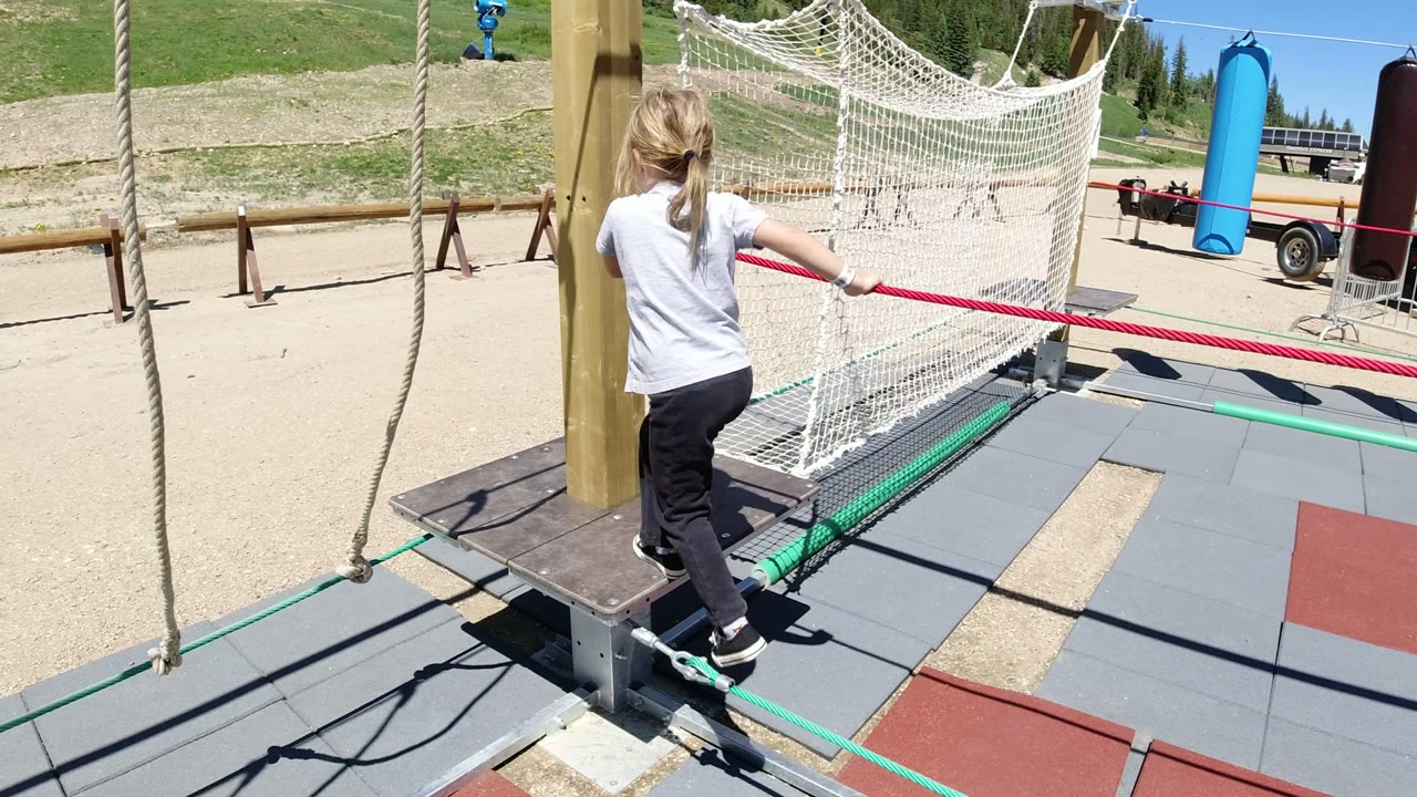 The girl steps forward near a wooden post, using the playground structure beside red rope and white netting in Winter Park, Colorado.
