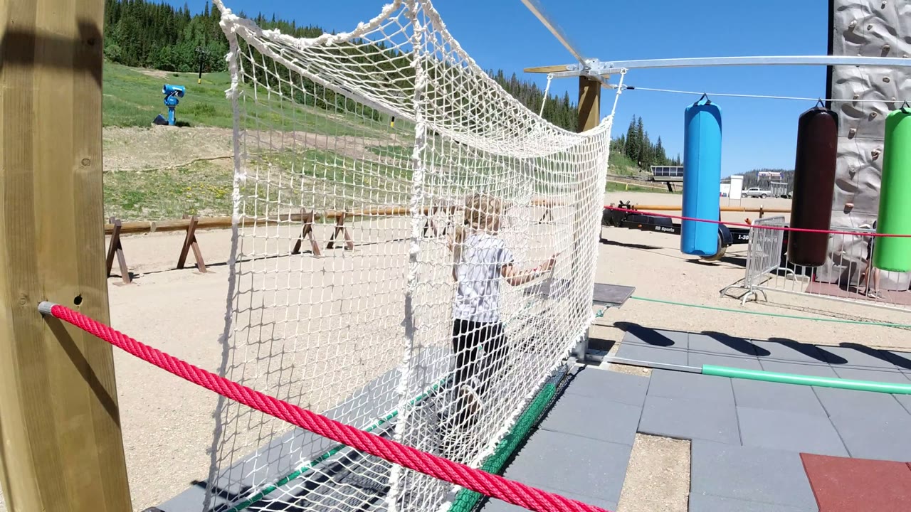 A child stands behind a white rope net at a Winter Park, Colorado playground, with sand, green hillside, and colored hanging obstacles in bright sun.