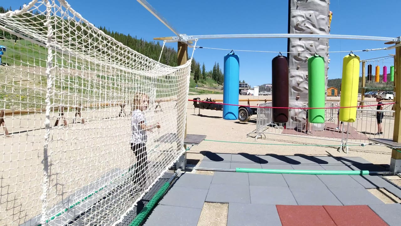 The child grips the rope net in the Winter Park playground, with gray mats below, a blue hanging obstacle, and activity-course equipment behind.