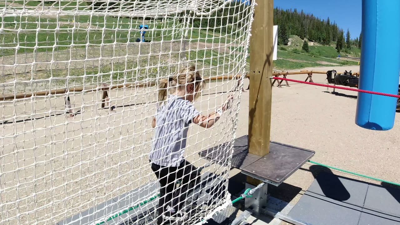 Girl holds the white net near a wooden post on the Winter Park playground, with blue play equipment, red rope, sandy ground, and green hillside behind her.
