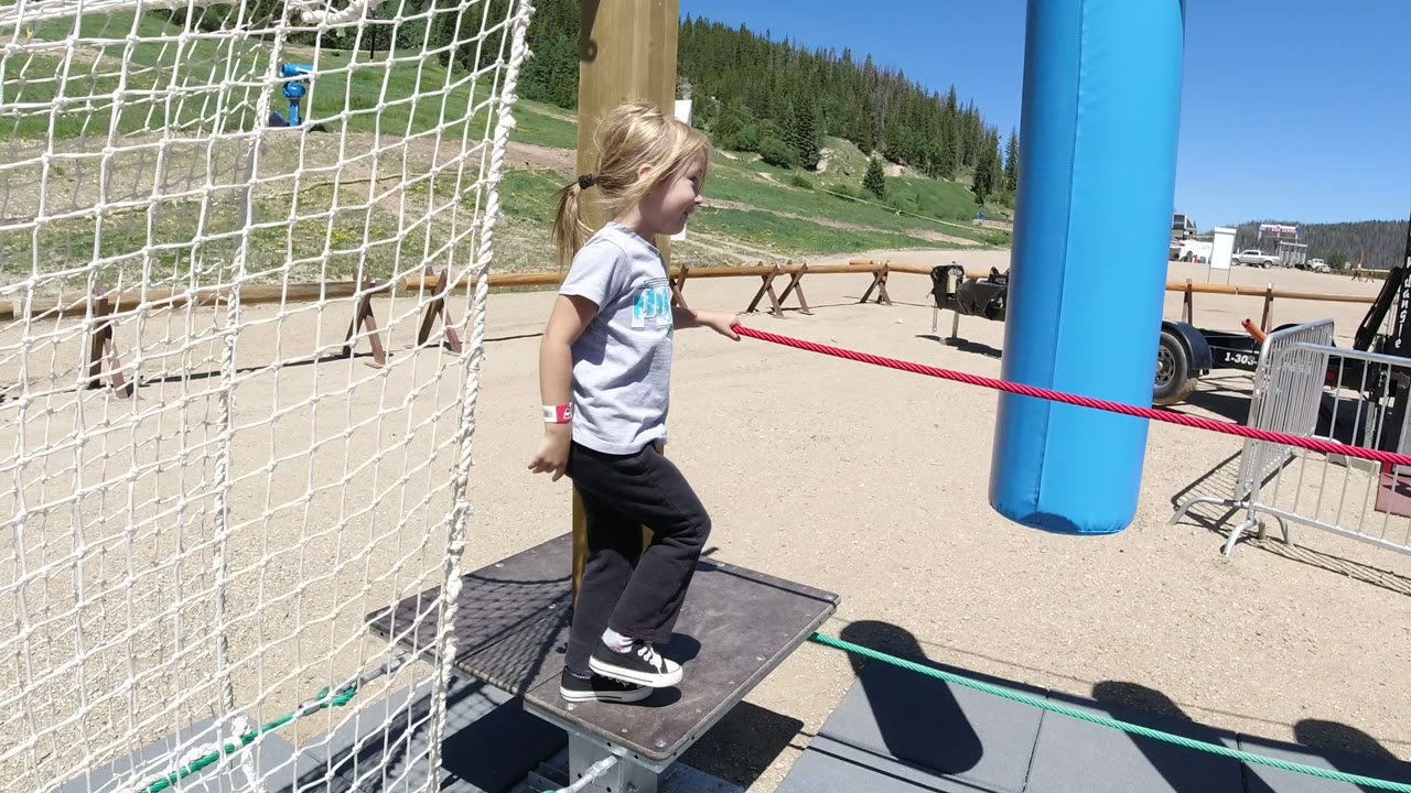 The girl stands on a small platform in Winter Park, Colorado while holding the red rope, with white netting at left and the blue hanging obstacle ahead of her.
