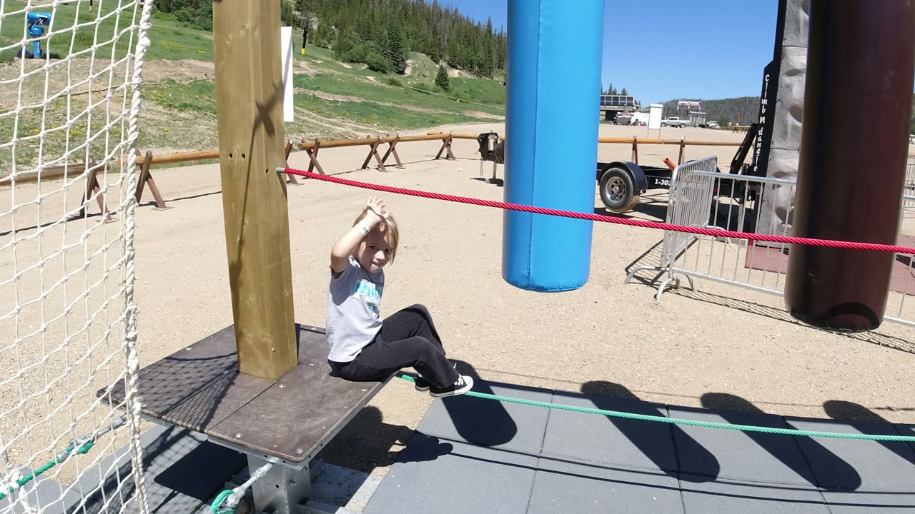 The child remains seated on the ropes course platform in Winter Park, Colorado, with her arms raised around the red rope and padded hanging obstacles beside her.